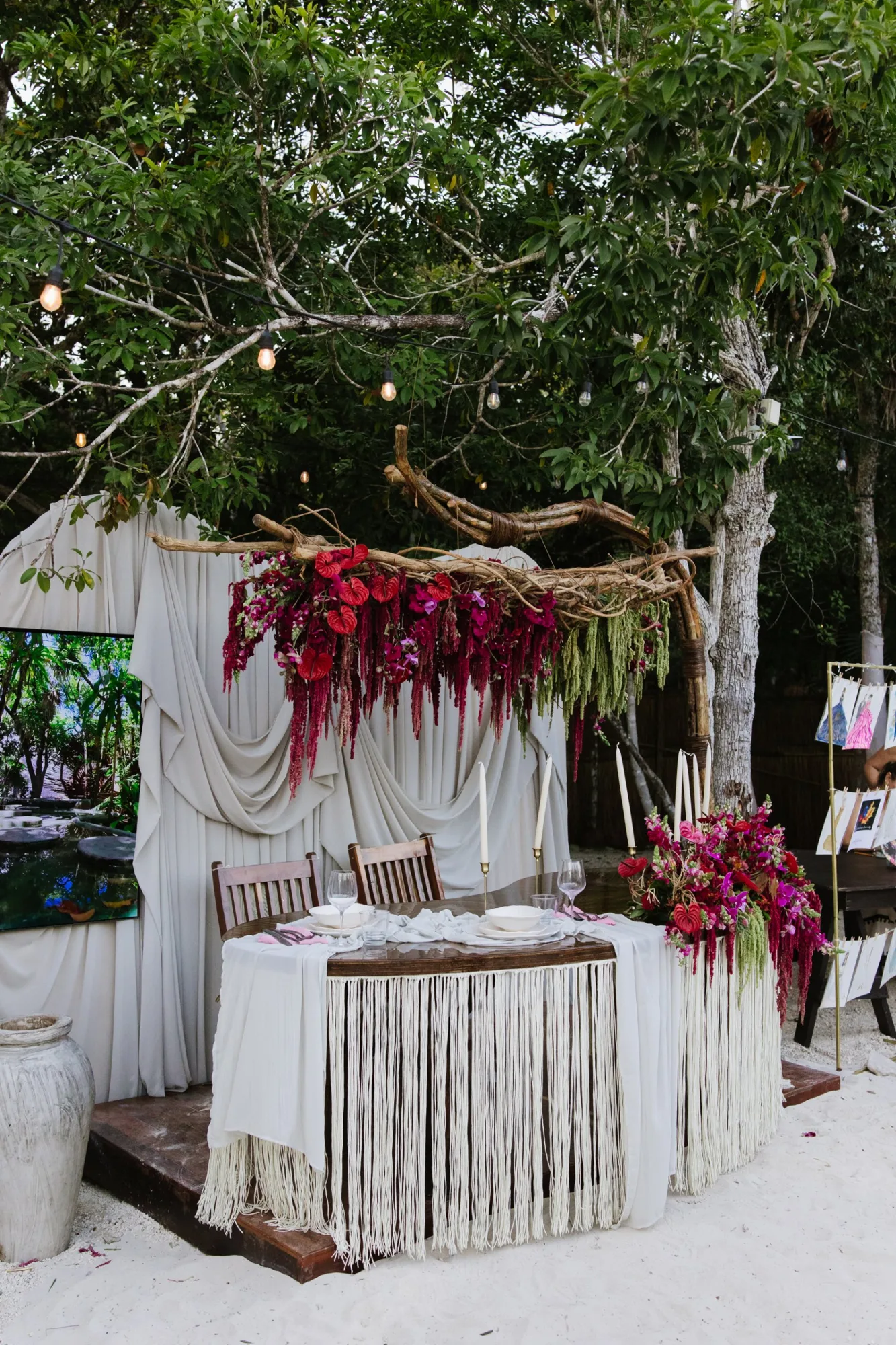 A sweetheart table set with white linens, macramé and red floral arrangements
