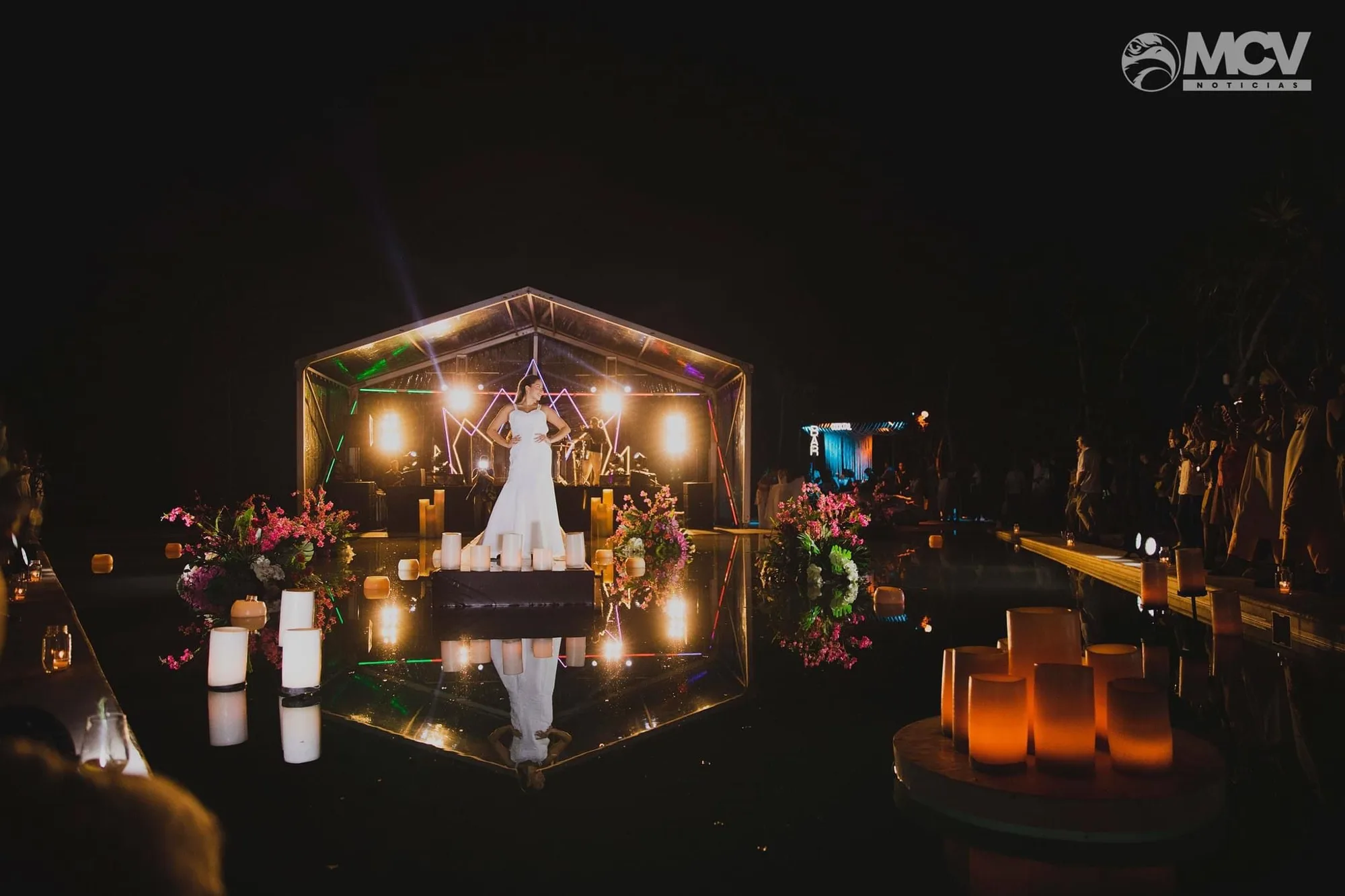 A bride stands on a mirror-like runway over water at night, with candles and flowers reflected beside a lit pavilion