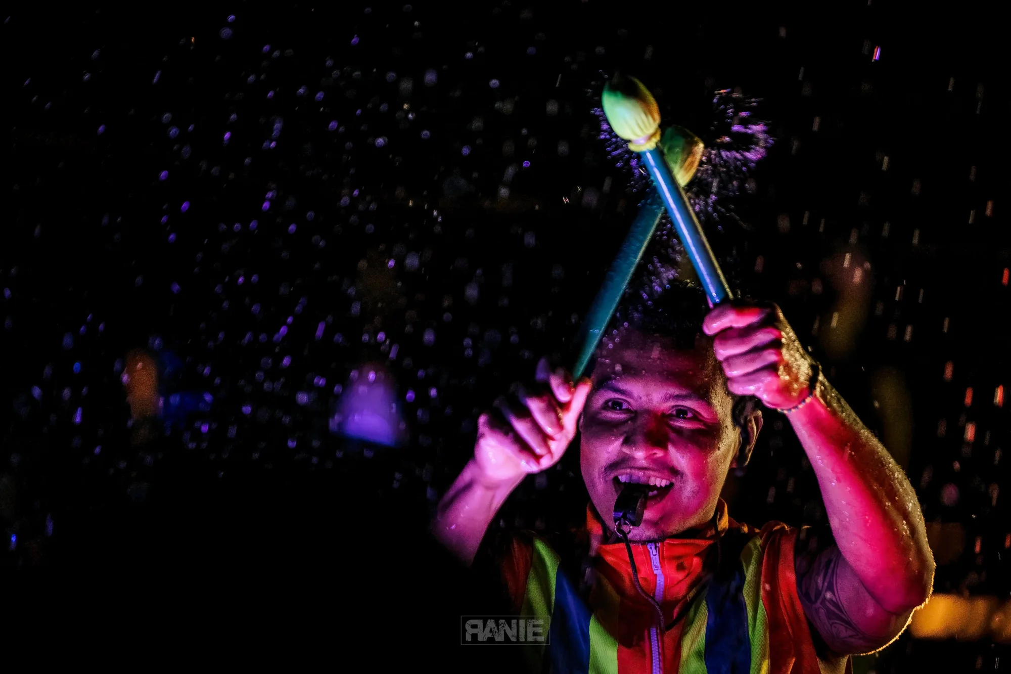 A percussionist in a colourful vest raises mallets through a spray of water droplets in the night air