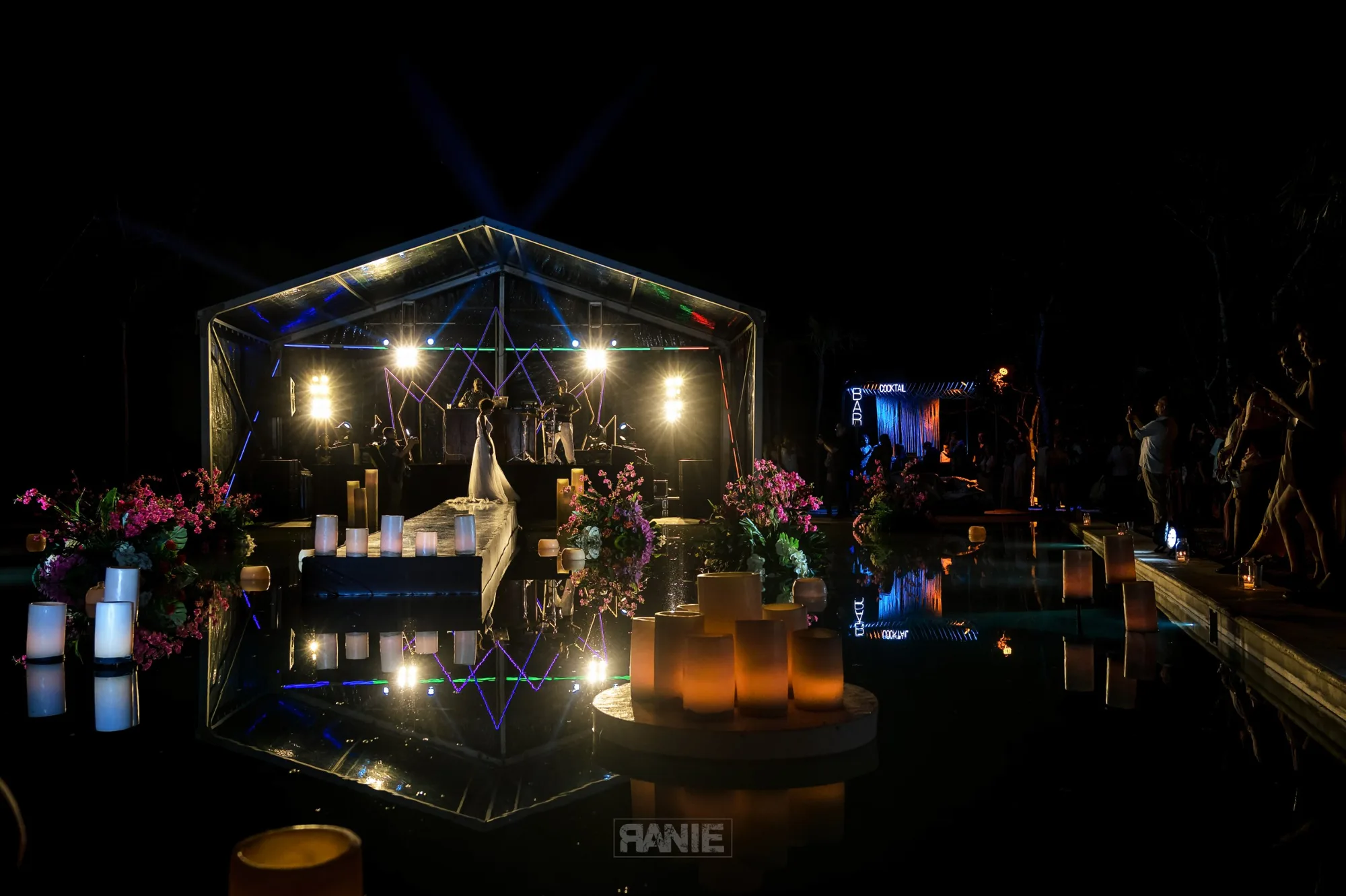 Floating candles and pink orchids line a mirrored runway over water leading to a glowing pavilion stage at night