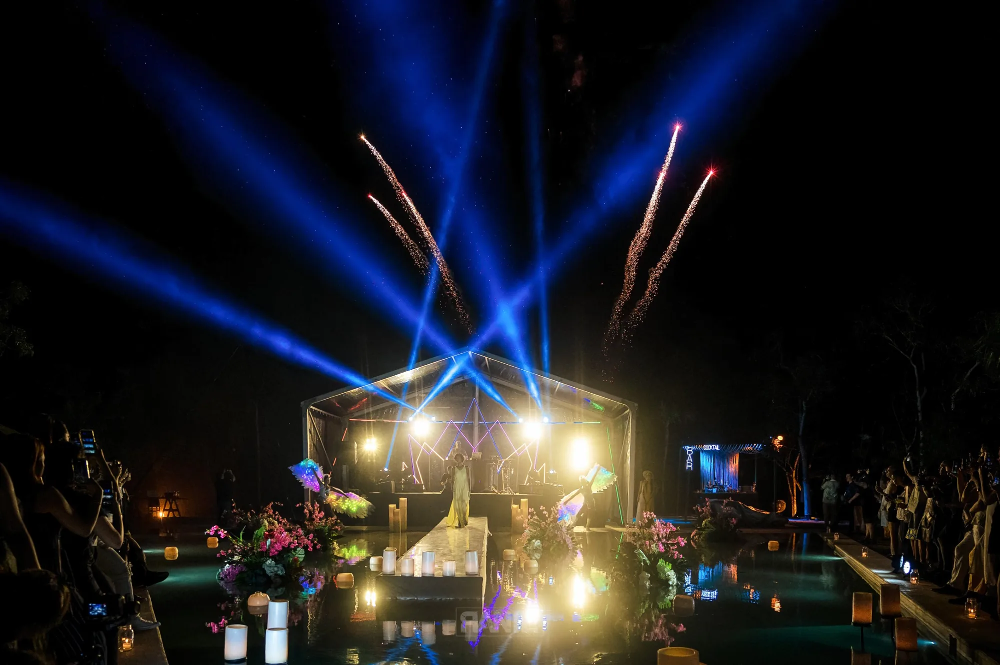Blue beams of light and fireworks burst over an illuminated stage above a pool lined with floating candles and flowers as guests watch from the edge