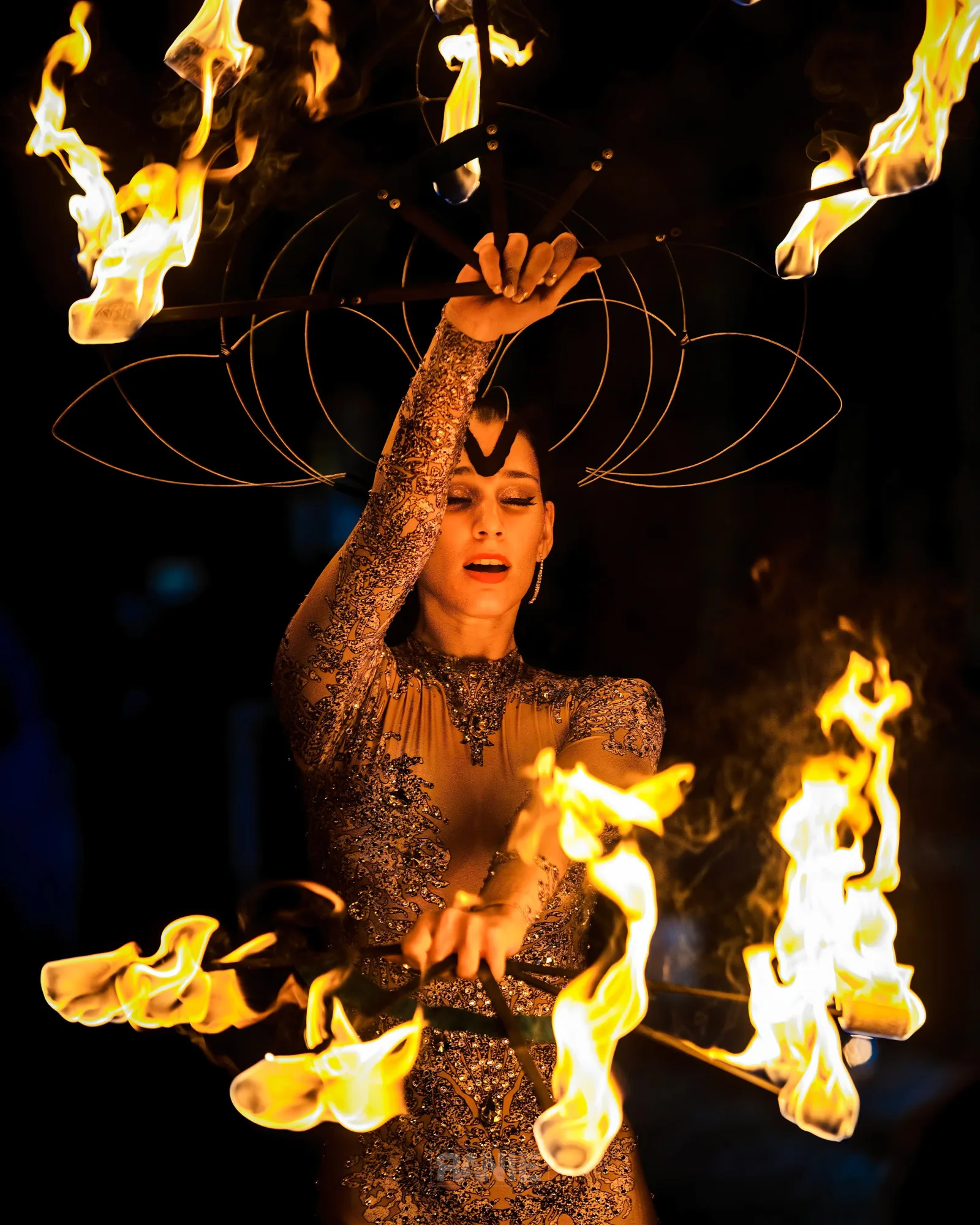 A fire performer in an embellished lace costume spins a flaming prop against the night sky