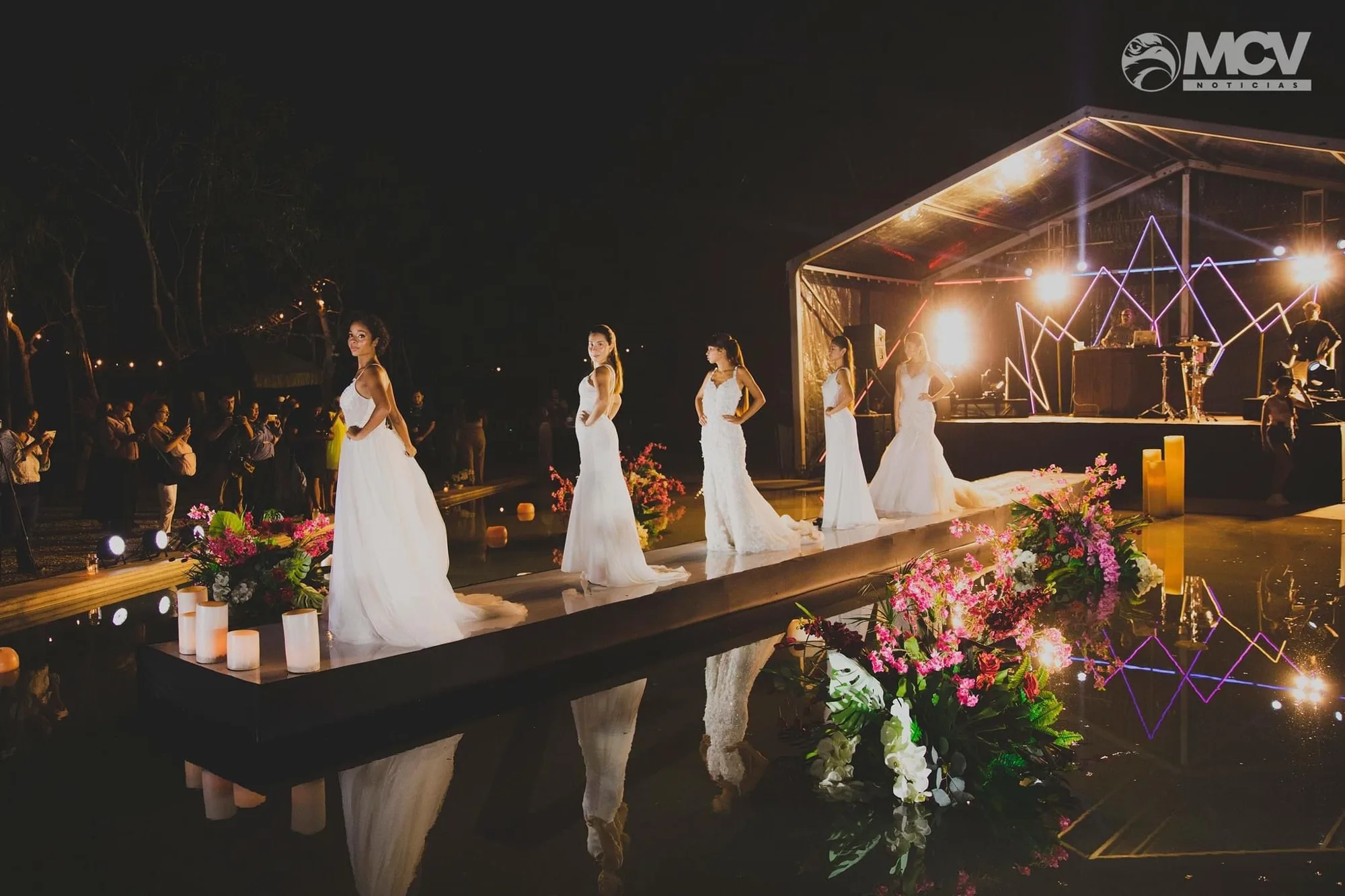 Brides in white gowns walk a mirrored runway over water beneath a glowing pavilion at night