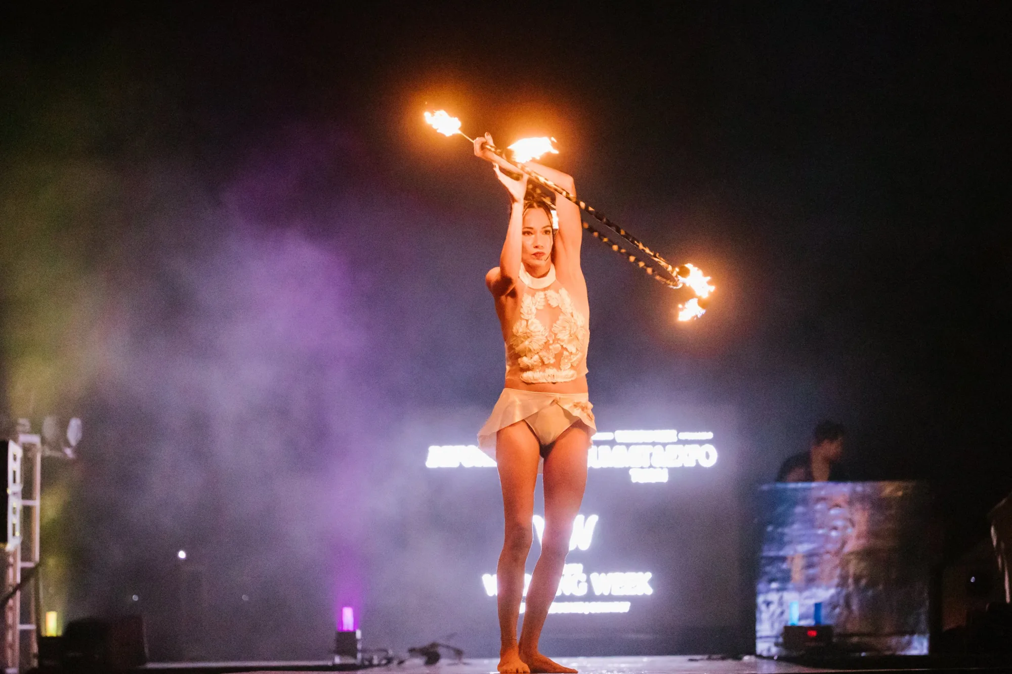 A fire performer spinning flaming staffs on stage through the smoke